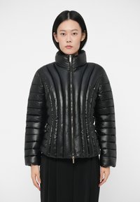 Svart polstret jakke med høy krage, glidelås og vertikal stripet tekstur. Laget av blankt materiale, med en tettsittende design.