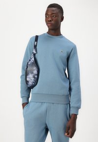Felpa azzurra con polsini e orlo a coste, dotata di un piccolo logo verde. Abbinata a pantaloni coordinati e una borsa a tracolla con effetto tie-dye scuro.