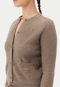 Stickad cardigan i ljusbrun färg med ett strukturerat diamantmönster, två knappar och en liten framficka. Mjuk, figurnära design.