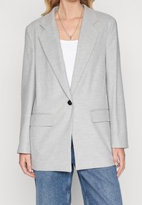 Blazer gris clair avec un bouton noir unique, des revers crantés et deux poches avant. Coupe ajustée, fabriqué dans un tissu texturé.