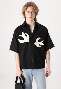 Camisa preta de manga curta com bordado de pássaro branco em contraste no peito, com um corte descontraído e fecho frente com botões.