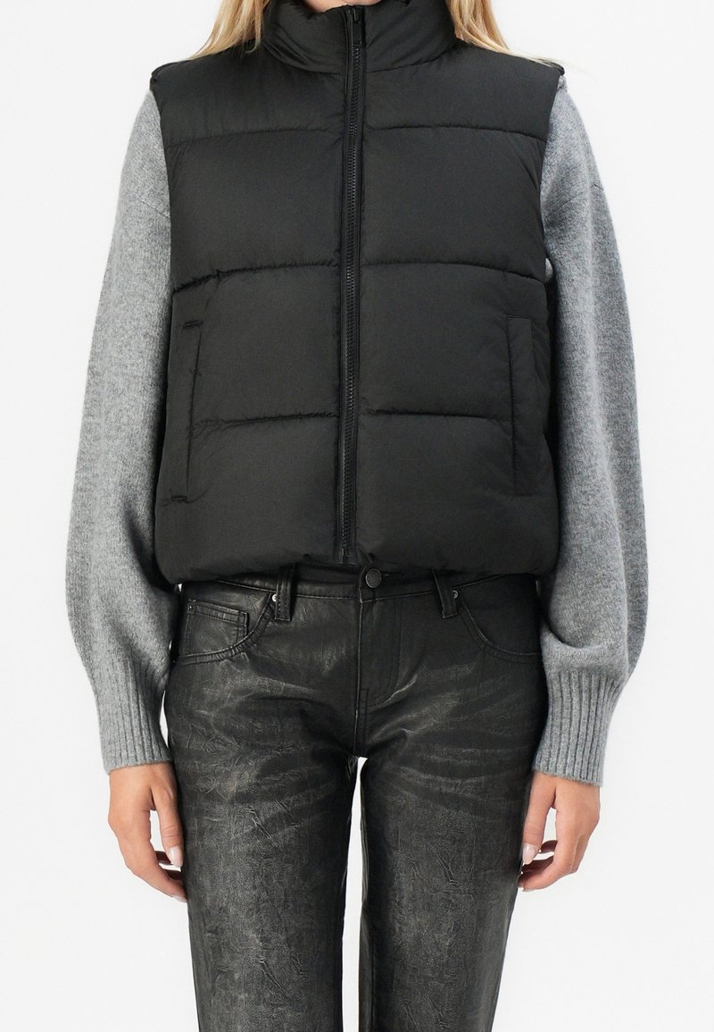 Personne portant une veste matelassée noire par-dessus un pull gris à manches longues avec un pantalon noir texturé, debout les mains détendues le long du corps.