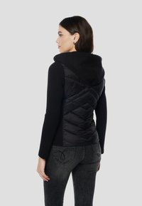 Črno dolgotrajno majico z dolgimi rokavi, podloženo s črno puhasto prešito telovnikom s kapuco. Gladek material z diamantnimi vzorci na telovniku. Oprijet kroj.