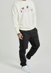 Man i vit sweatshirt med fjäril och "Siksilk"-text, svarta cargobyxor och vita sneakers, står mot en enfärgad bakgrund.