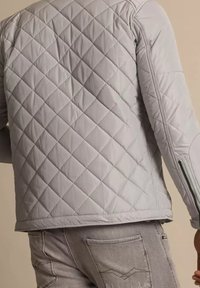 Giacca leggera grigia trapuntata con motivo a rombi indossata sopra jeans grigi sbiaditi, caratterizzata da una cerniera sul manica e cuciture dettagliate sui pantaloni.