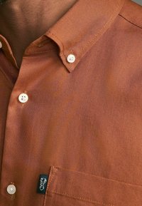 Braunes Hemd zum Knöpfen aus strukturiertem Stoff, mit weißen Knöpfen, einer Brusttasche und einem kleinen schwarzen Logotag an der Seite.