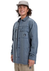 Hellblaues Jeansjackett mit Knopfverschluss, zwei Brusttaschen und einem Logopatch. Lange Ärmel und lässiger Schnitt, getragen über beigen Hosen.
