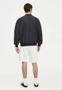 Sort bomberjakke med ribbede manchetter og krave, parret med hvide stribede cargoshorts. Model vendt væk, iført sorte sneakers.