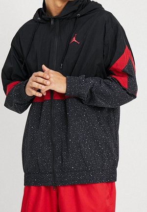 Person trägt eine schwarz-rot gefleckte Jacke mit Reißverschluss und Kapuze sowie rote Shorts, Hände vor dem Körper verschränkt.