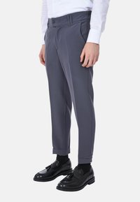 Pantaloni grigi con una texture liscia, caratterizzati da un taglio su misura, pieghe doppie e passanti per cintura, abbinati a scarpe e calze nere.