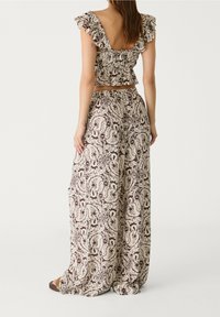 Vrouw in bijpassende paisley-print, volants crop top met open rug en wijde broek, staande tegen een effen witte achtergrond.