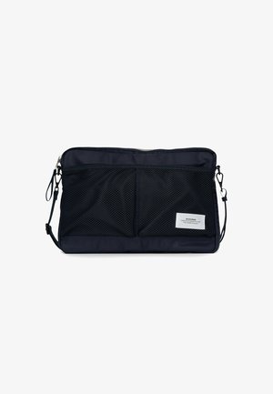 La bolsa de color azul marino tiene un cuerpo de tela suave, bolsillos frontales de malla, cremallera plateada y una correa desmontable. Presenta una etiqueta de marca en el costado.