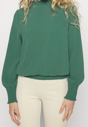 Frau trägt eine grüne Bluse mit langen Ärmeln und gerafften Bündchen sowie beige Hose, stehend vor einem einfarbigen Hintergrund.