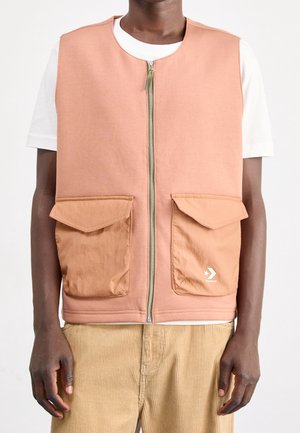 Chaleco melocotón sin mangas con cremallera frontal y dos grandes bolsillos de solapa, llevado sobre una camisa blanca de manga corta y pantalones beige de pana.