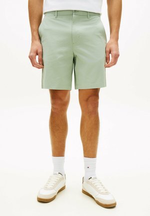 Mann trägt hellgrüne, maßgeschneiderte Shorts, weiße Kniestrümpfe und weiße Turnschuhe mit beigefarbenen Akzenten, steht auf weißem Hintergrund.