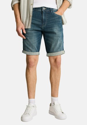 Man wearing rolled-up blue denim shorts, white sneakers, white socks, and a light striped shirt with one hand in pocket.