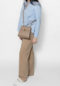 Borsa a tracolla in pelle beige con hardware dorato, forma rettangolare e chiusura a patta, indossata con una camicia azzurro chiaro e pantaloni beige a righe.