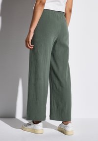 Pantaloni palazzo realizzati in tessuto verde testurizzato, con vita elasticizzata. Indossati con sneakers bianche, su uno sfondo grigio chiaro.