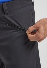 I pantaloni neri presentano una tasca laterale con zip. Il tessuto appare liscio con una leggera lucentezza, evidenziando una vestibilità sartoriale e un design pratico.