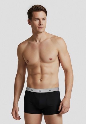 Skjorteløs mand med kort brunt hår iført sorte boxershorts med grå linning, stående mod en ensfarvet lys grå baggrund.