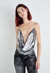 Top argento con paillettes, scollatura profonda a V e spalline sottili, abbinato a jeans grigi scuri strappati. Texture liscia e superficie riflettente.
