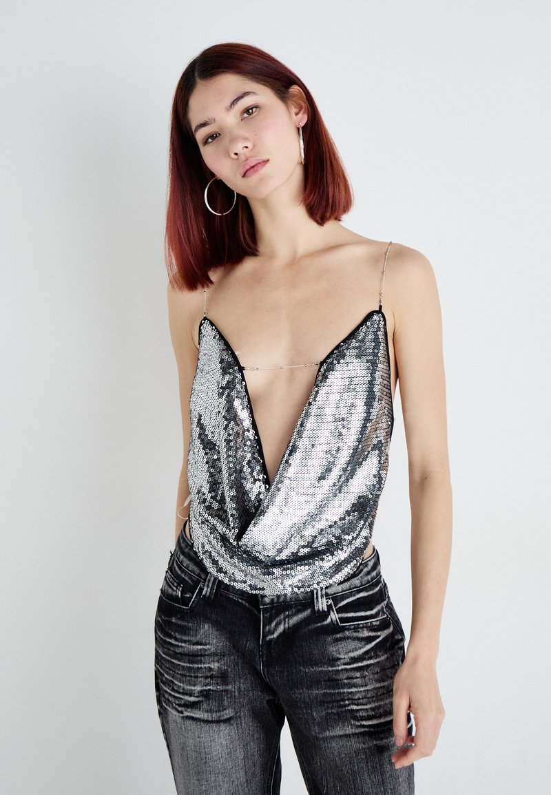 Top argento con paillettes, scollatura profonda a V e spalline sottili, abbinato a jeans grigi scuri strappati. Texture liscia e superficie riflettente.