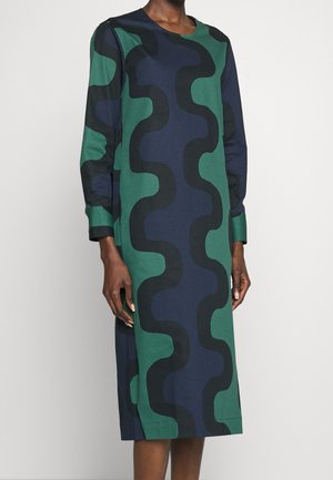 Frau trägt ein langärmeliges, knielanges Kleid mit grünen, marineblauen und schwarzen wellenförmigen abstrakten Mustern.