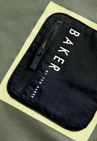 Donkergroene stof met een zwarte zak met rits. Witte letters tonen "BAKER by TED BAKER" op de voorkant van de zak.