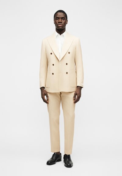 Homme portant un costume croisé beige avec un pantalon assorti, une chemise blanche et des chaussures habillées noires, debout devant un fond uni.