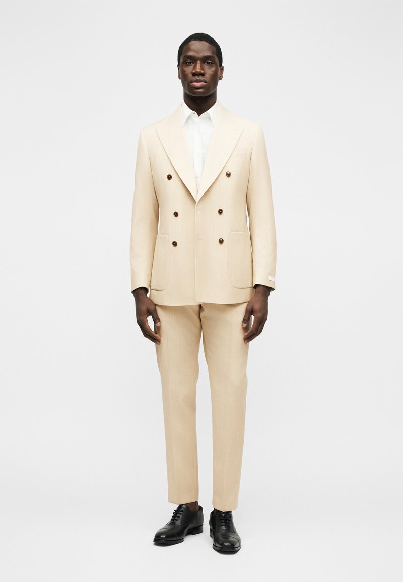 Άνδρας ντυμένος με μπεζ κοστούμι διπλής σειράς κουμπιών και ταιριαστό παντελόνι, λευκό πουκάμισο και μαύρα επίσημα παπούτσια, στέκεται μπροστά σε απλό φόντο.