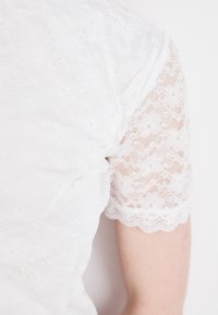 Hvit topp med korte blondeermet med blomstermønster og bølgete kanter, som komplementerer en glatt hvit stoffkropp.