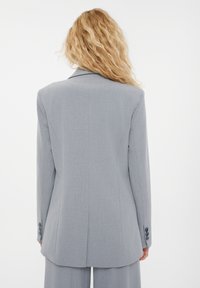 Blazer grigio su misura con uno spacco singolo, rever a scollatura e polsini a quattro bottoni. Realizzato in tessuto testurizzato con una silhouette strutturata.