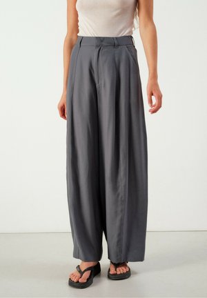 Kvinde iført højtaljede, vidde mørkegrå bukser kombineret med en ærmeløs beige top og sorte sandaler, stående mod en ensfarvet baggrund.