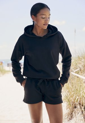 Schwarzer Baumwollhoodie mit einer Kordelzugkapuze, kombiniert mit schwarzen Shorts mit einem elastischen Bund, glatter Textur und lockerer Passform.