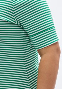 Zöld-fehér csíkos rövid ujjú ing puha, sima anyagból; testhez álló fazon és felhajtott mandzsetták, amelyek kiemelik a merész mintát.