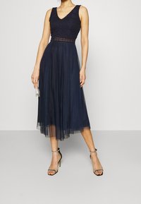 Abito da sera blu navy con un corpetto in pizzo, scollatura a V e una gonna in tulle a strati. Presenta una vita aderente e include tacchi neri con cinturini.