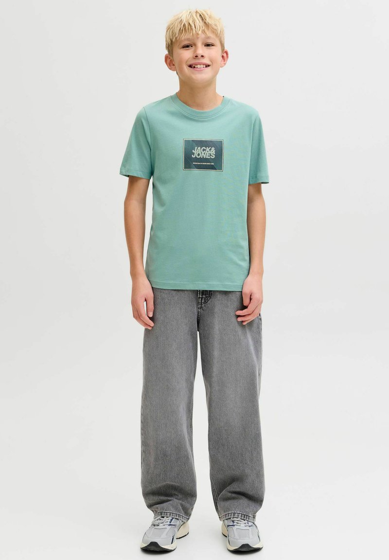 Mintgroen katoenen T-shirt met een rechthoekig "JACK & JONES" logo, gecombineerd met losse grijze spijkerbroek en grijze sneakers.