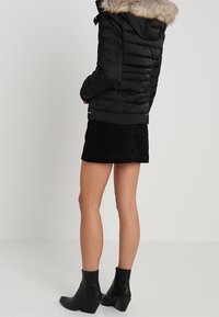 Czarna kurtka puchowa z kapturem wyłożonym futrem, z pikowanym wzorem i ribbowanymi detalami, noszona na czarnej spódnicy i czarnych botkach za kostkę.