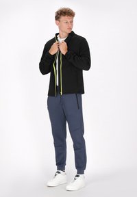 Zwarte zip-up jack met een groene rits, gedragen over een wit shirt, gecombineerd met blauwe joggingsbroeken en witte sneakers. Voorzien van ritszakken.