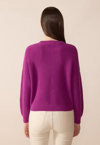 Fuchsia strikkegenser med rund hals, senkede skuldre og ribbestrikket tekstur. Kombinert med hvite tettsittende jeans for kontrast.