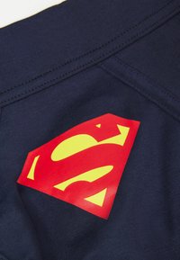 Tissu bleu marine avec une texture lisse, arborant un logo Superman rouge et jaune bien visible. Comprend des détails cousus le long des coutures.