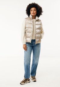 Ljusbeige puffer-väst med hög krage och kviltad design, bärs över en krämfärgad tröja, parat med blå jeans och bruna sneakers.