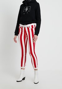 Jeans a rayas rojas y blancas con un diseño ajustado, combinados con una sudadera negra con capucha y botas blancas hasta el tobillo. Cinturón blanco incluido.