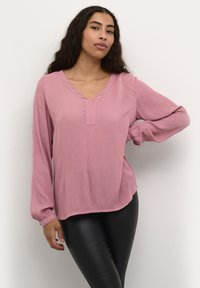 Langärmlige Bluse mit V-Ausschnitt in sanftem Rosa und subtiler Textur, mit geradem Saum und lässiger Passform. Kombiniert mit schwarzen Lederleggings.
