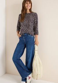 Mönstrad blus med långa ärmar i mörk lila, kombinerad med vida blå jeans. Håller en krämfärgad axelväska; vita sneakers med röda detaljer.
