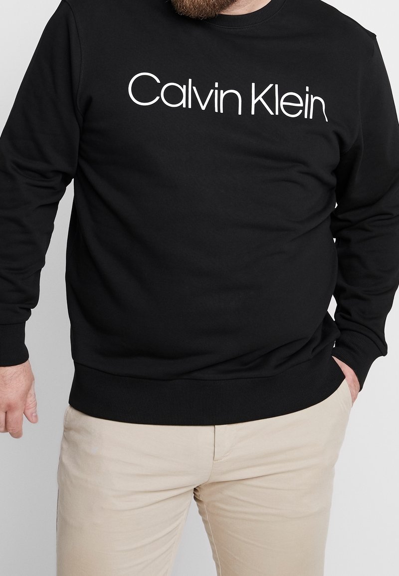 Mustat puuvillaneuleet, joissa on pyöreä kaula-auko, resorit hihansuissa ja helmassa. Etupuolella valkoisella painettuna "Calvin Klein" -logo.