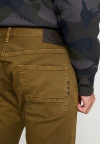 Widok z tyłu osoby w brązowych spodniach z skórzaną naszywką marki oraz ciemnej koszuli z długim rękawem w kamuflażu.