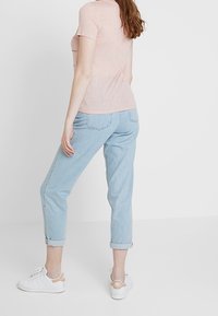 Jasnoróżowy t-shirt z krótkim rękawem, w zestawieniu z jasnoniebieskimi dżinsami z podwiniętymi nogawkami oraz białymi trampkami z beżowymi akcentami, widziany od tyłu.