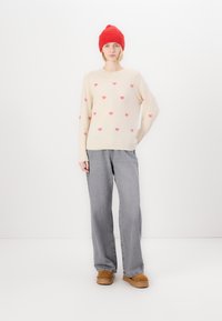 Pull beige avec des motifs de cœurs roses, associé à un pantalon large gris et des bottes tan. Le mannequin porte un bonnet en tricot rouge. Fond minimaliste.