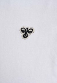 Hvidt stof t-shirt med et lille broderet sort bi-logo. Sømmene er detaljeret med en beige kant omkring bien.